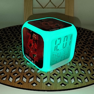 Op de afbeelding: Een kubusvormige digitale wekker met een gloeiend turquoise frame. De klok geeft de tijd, datum en temperatuur in groen weer. Eén kant heeft een foto van een voetballer in een rood uniform. Het woord "TIMEO" staat op de klok.