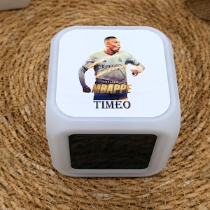 Op de afbeelding: Witte digitale wekker met een zwart display. De voorkant van de wekker heeft een geprinte afbeelding van een voetballer in een wit shirt met de naam "Kylian Mbappé" en het woord "Timeo" eronder.