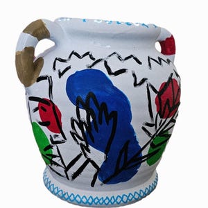 Może przedstawiać: Biały ceramiczny wazon z dwoma uchwytami, pomalowany abstrakcyjnymi wzorami w kolorze niebieskim, czerwonym i zielonym. Wazon ma niebiesko-białą wzorzystą krawędź.
