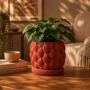 May include: A vibrant terracotta-coloured plant pot featuring a repeating vertical leaf-like texture pattern. The cylindrical pot sits on a matching circular base and contains a lush green plant with heart-shaped leaves, creating a distinct visual contrast.