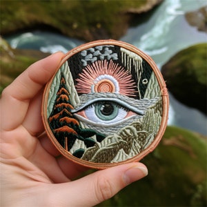 Könnte beinhalten: Gestickter Patch mit einem farbenfrohen Auge, umgeben von einer Landschaftsszene. Das Auge ist blau und grün mit einer weißen Iris. Die Landschaft ist eine Mischung aus Grün, Braun und Rosa mit einer aufgehenden Sonne im Hintergrund.
