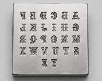 May include: A square silver-toned metal plate featuring a debossed alphabet from A to Z. The letters are arranged in five rows, with each character displaying a decorative serif font style. The surface of the metal plate has a brushed finish.
