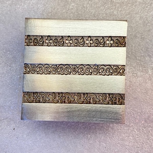 May include: A square, silver-colored metal object with three horizontal bands of ornate, dark brown floral designs. The bands are separated by wider, brushed silver sections. The object has a simple, elegant design.
