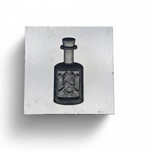 May include: A dark glass bottle with a cylindrical body and a small, round top. The bottle features a label with a skull and crossbones design. The bottle is set on a square, light-colored surface.