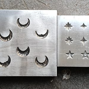 Può includere: Due timbri in metallo argentato per la creazione di gioielli. Un timbro presenta sei forme di luna crescente con dettagli decorativi. L'altro timbro presenta nove forme di stella e una lettera "C".