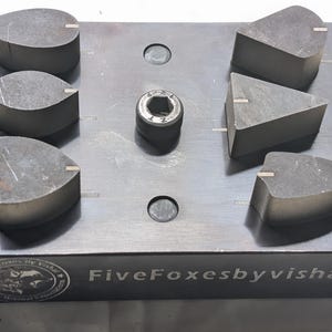 Puede incluir: Un bloque de metal gris oscuro con seis piezas de piedra grises de formas únicas y un perno central plateado. El bloque tiene forma rectangular con esquinas redondeadas. El texto "Five Foxes by Vishal" está impreso en el lateral.