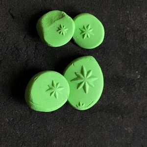 May include: Four green, circular and oval-shaped clay stamps with starburst designs. The stamps are arranged on a dark, textured surface. The stamps vary in size and shape, with the starbursts pressed into the clay.