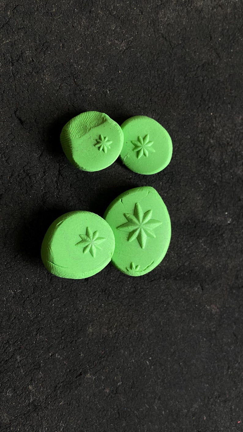 May include: Four green, circular and oval-shaped clay stamps with starburst designs. The stamps are arranged on a dark, textured surface. The stamps vary in size and shape, with the starbursts pressed into the clay.