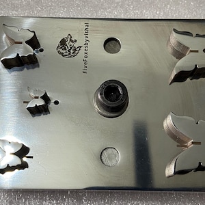 Puede incluir: Placa rectangular de metal cepillado con cinco recortes de mariposas y un perno central. La placa tiene un pequeño logotipo de zorro y el texto "FiveFoxesbyVishal". También hay dos agujeros circulares.