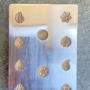 Op de afbeelding: Een rechthoekige, geborstelde metalen plaat met diverse reliëf, goudkleurige bloemen- en paisley-ontwerpen. De ontwerpen omvatten een druppel, een schelp, een bloem en een paisley-vorm. De plaat heeft een zilveren afwerking.