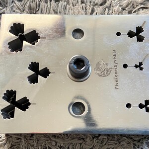 May include: A rectangular, silver-colored metal object with several cut-out shapes resembling stylized flowers. It has a central, dark-colored cylindrical component and two circular holes. The text "FiveFoxesbyVishal" is visible on the surface.