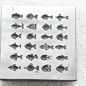 May include: A square, silver-colored metal stamp with a grid of 24 small fish designs. Each fish is a different style, with varying fins, tails, and body shapes. The stamp is likely used for imprinting designs onto materials like clay or metal.