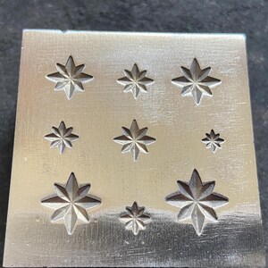 May include: A silver metal stamp with nine star-shaped designs of varying sizes. The stars are evenly spaced on the rectangular surface. The stamp is likely used for jewelry making or other crafts.