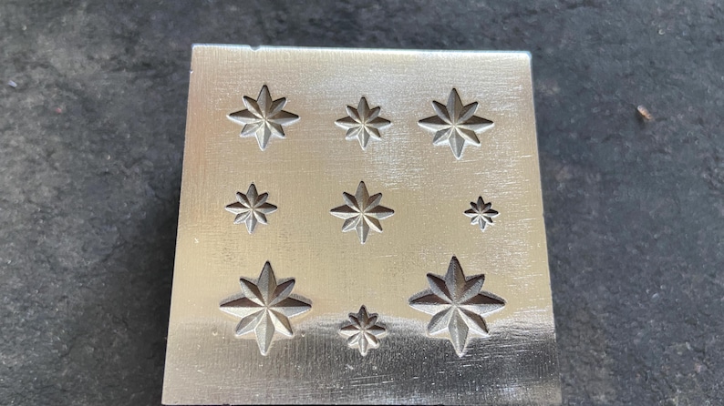 May include: A silver metal stamp with nine star-shaped designs of varying sizes. The stars are evenly spaced on the rectangular surface. The stamp is likely used for jewelry making or other crafts.