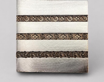 May include: A square metal object with a brushed silver finish featuring three horizontal bands of textured, dark bronze-coloured decorative patterns. The design consists of alternating smooth silver strips and intricate, embossed floral and scrollwork.