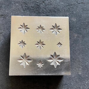 May include: A square, silver-colored metal stamp with nine star-shaped impressions of varying sizes. The stars have eight points and are evenly spaced across the surface. The stamp is likely used for jewelry making or other crafts.