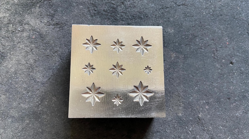 May include: A square, silver-colored metal stamp with nine star-shaped impressions of varying sizes. The stars have eight points and are evenly spaced across the surface. The stamp is likely used for jewelry making or other crafts.
