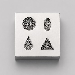 May include: A square silver-toned metal block featuring four recessed impressions with intricate starburst patterns. The shapes include a circle, an oval, a teardrop, and a triangle, each centered with a small dot and radiating lines.
