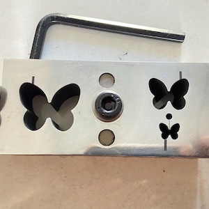 Può includere: Un utensile per fustellatura a farfalla in metallo con finitura argentata. L'utensile presenta ritagli a farfalla e forme di farfalla attaccate. Una chiave a brugola è appoggiata sopra l'utensile.