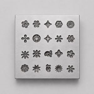 May include: A square silver-coloured metal block featuring a grid of twenty recessed impressions. The impressions consist of various floral, star, and abstract geometric shapes, each etched into the surface of the metal plate in a four-by-five arrangement.