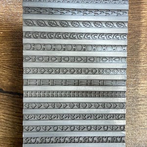 May include: A rectangular metal stamp with multiple rows of decorative border designs. The borders feature various patterns, including leaves, circles, and geometric shapes. The stamp is grey and appears to be used for creating embossed designs.