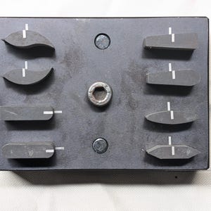 Puede incluir: Una placa metálica gris oscuro con ocho piezas de goma negra de varias formas, cada una con una línea blanca. La placa tiene un agujero central y dos tornillos. El fondo es una superficie blanca.