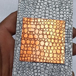 Puede incluir: Una pieza rectangular de metal con un patrón texturizado gris, similar a burbujas. Un cuadrado de metal color cobre con un patrón similar está insertado. La pieza se sostiene en una mano.