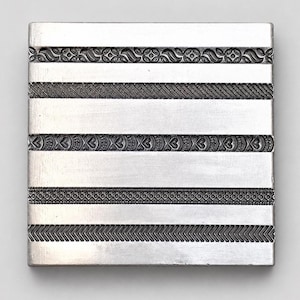 May include: A square silver-toned metal stamping plate featuring five distinct horizontal bands of embossed patterns, including floral motifs, diagonal lines, heart shapes, a fine dotted texture, and a repeating chevron design.
