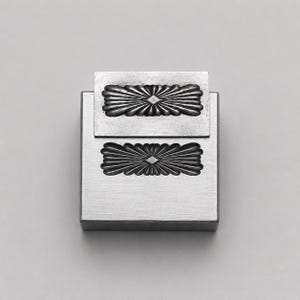 Könnte beinhalten: Ein rechteckiger silberfarbener Metallstempel mit einem erhabenen, länglichen, blumenartigen Motiv und einer zentralen Rautenform. Das Design wiederholt sich sowohl auf dem Hauptblock als auch auf dem kleineren, leicht versetzten Aufsatz.