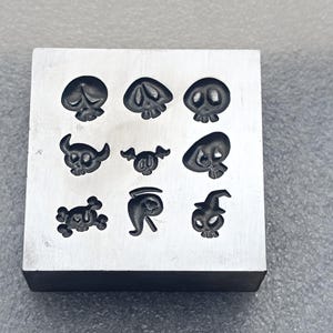 May include: A square metal stamp block with nine skull designs. The skulls vary in style, some with hats and others with crossbones. The block is silver and sits on a gray surface, perfect for crafting Halloween-themed items.