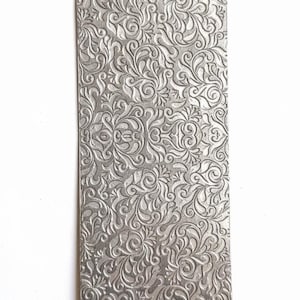 May include: A rectangular, silver-coloured metal plate with an embossed floral pattern. The design features swirling vines and leaves, creating a textured surface. The plate is set against a white background.