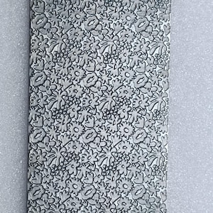 Könnte beinhalten: Rechteckige Metallplatte mit detailliertem Blumenmuster. Das Design zeigt eine durchgehende Anordnung stilisierter Blumen, Blätter und anderer organischer Formen, wodurch eine strukturierte Oberfläche entsteht. Die Platte ist neutralgrau.