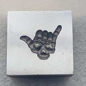 May include: A square, silver-colored metal block with a detailed engraving of a skull-shaped hand gesture. The design features a skull face within the hand, with the thumb and pinky extended. The block has a brushed metal finish.