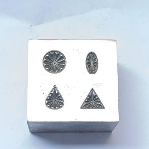 May include: A silver metal block with four different shaped impressions: a circle, an oval, a teardrop, and a triangle. Each shape has a detailed, radiating design. The block sits on a white surface.