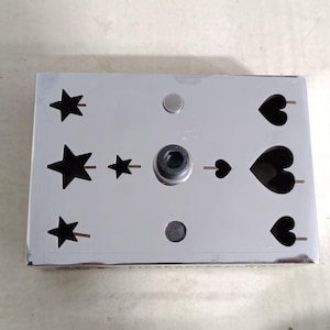 Puede incluir: Una herramienta de corte de metal plateado con formas de estrellas y corazones. La herramienta tiene un mecanismo central y está diseñada para manualidades o fines decorativos. Las formas troqueladas están espaciadas uniformemente.