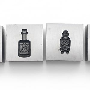 May include: Four square metal blocks with engraved designs. The designs include a hand making the rock and roll sign with a skull, a bottle, a bottle with a skull and crossbones, and a hand making the shaka sign with a skull.