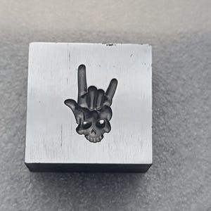 May include: A square, silver-colored metal block with a carved design. The design is a hand gesture with the index and pinky fingers extended, forming a rock-and-roll symbol, with a skull in the center. The block is set against a gray background.