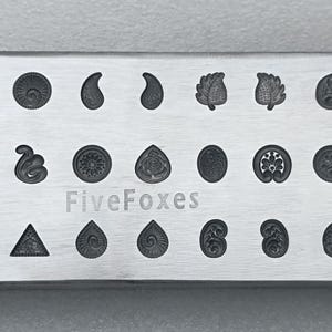 May include: A rectangular metal stamp block featuring an array of decorative designs. These include paisley shapes, leaves, and circular patterns. The text "Five Foxes" is etched onto the block.