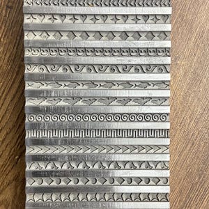 May include: A rectangular metal stamp with various engraved border designs. The stamp features rows of repeating patterns, including leaves, hearts, and geometric shapes. The metal has a silver color and a textured surface.