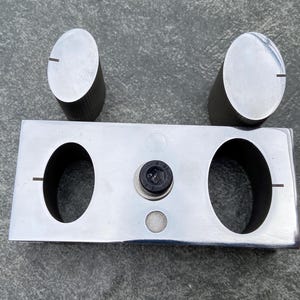 May include: A silver-colored metal object with two oval-shaped knobs and a rectangular base. The base has two large oval cutouts and a central circular element. Each knob has a single, thin groove.