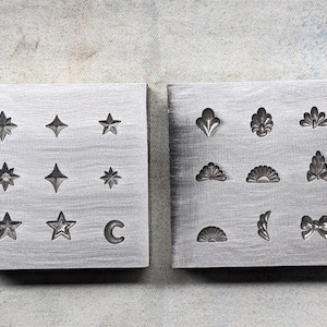 May include: Two square metal blocks with various star, moon, and decorative shapes. The left block has star and crescent moon designs. The right block features floral and bow shapes. The blocks are on a textured surface.