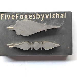 May include: Two metal clock hands, one with a pointed end and the other with a decorative design, are displayed on a dark gray rectangular base. The text "FiveFoxesbyvishal" is printed in white on the base.
