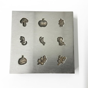 Könnte beinhalten: Eine Sammlung von neun kleinen Metallanhängern mit verschiedenen von der Natur inspirierten Designs, darunter ein Pilz, ein Apfel und eine Eidechse, angeordnet auf einer quadratischen, gebürsteten Metalloberfläche. Die Anhänger sind in Bronze- und Silbertönen gehalten.