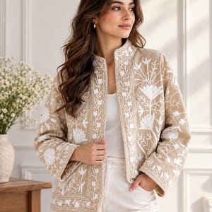 May include: A beige open-front jacket featuring intricate white floral embroidery throughout the fabric. The garment has a mandarin collar, long sleeves with wide cuffs, and a straight hemline, worn over a plain white top and white trousers.