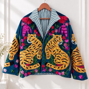 Puede incluir: Una chaqueta de color verde azulado con bordados de tigres amarillos vibrantes en los paneles frontales y las mangas. El diseño incluye detalles florales en rosa y rojo. La chaqueta tiene un cuello con solapa y un forro de rayas verticales blancas y verde azulado.