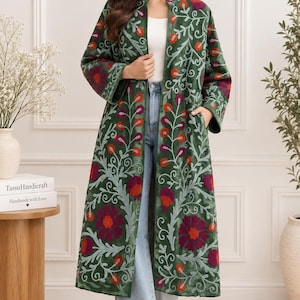 May include: A long, dark green coat featuring intricate floral embroidery in shades of red, purple, and light blue. The coat has a stand-up collar, long sleeves, and an open front design, worn over a white top and light blue denim jeans.