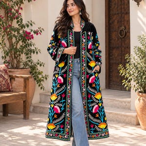 May include: A long black coat featuring vibrant, multi-coloured floral embroidery in shades of pink, yellow, and blue. The garment has a mandarin collar, long sleeves, and a straight-cut silhouette, worn over a black top and light blue denim jeans.