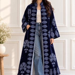 May include: A long, navy blue velvet duster coat featuring light blue embroidered circular and floral patterns along the front panels and sleeves. The coat has a stand-up collar, long sleeves, and side pockets, worn over a white top and blue jeans.