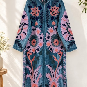 May include: A long-sleeved, zip-front coat in blue velvet, featuring intricate embroidery in shades of pink, dark blue, and coral. The design showcases large floral and botanical motifs that extend from the collar down to the hemline.