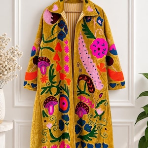 Pode incluir: Um casaco de manga comprida amarelo-mostarda com bordados vibrantes e multicoloridos de flores e cogumelos. O casaco tem uma gola, uma frente aberta e um forro às riscas visível na gola. O bordado inclui fios cor-de-rosa, azuis, verdes e cor de laranja.